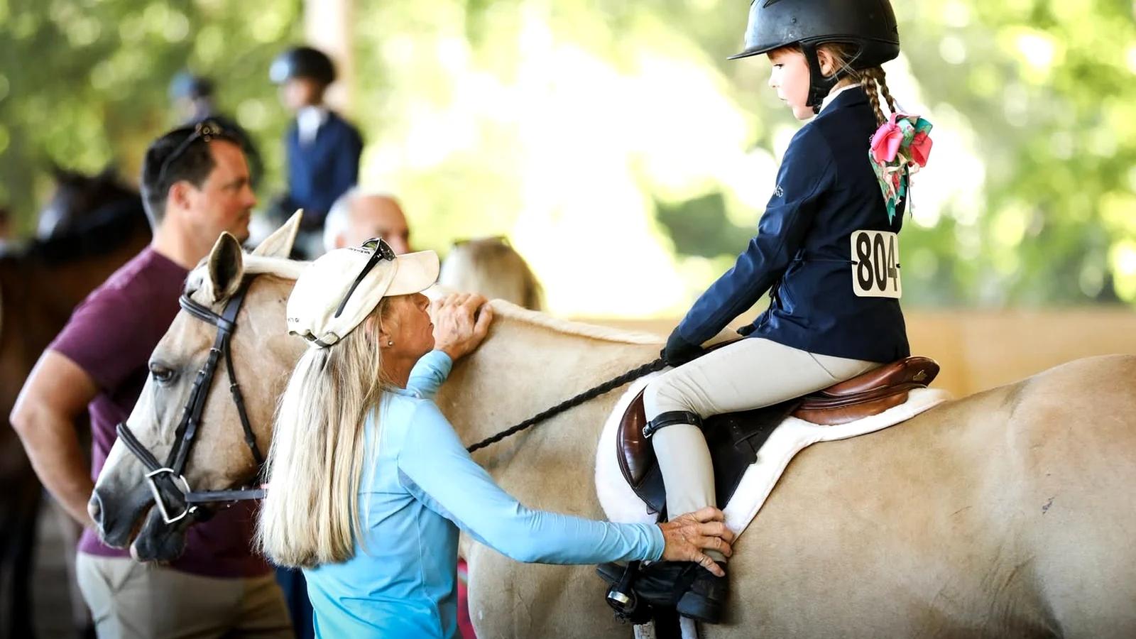 Instructor helping a young rider seated on a light-colored pony during a lesson.